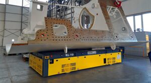 Componente meccanico di grandi dimensioni sostenuto da un veicolo automatizzato di trasporto giallo e blu con la scritta 'MORELLO' e il simbolo di capacità 'WLL 110t', all'interno di un capannone industriale.